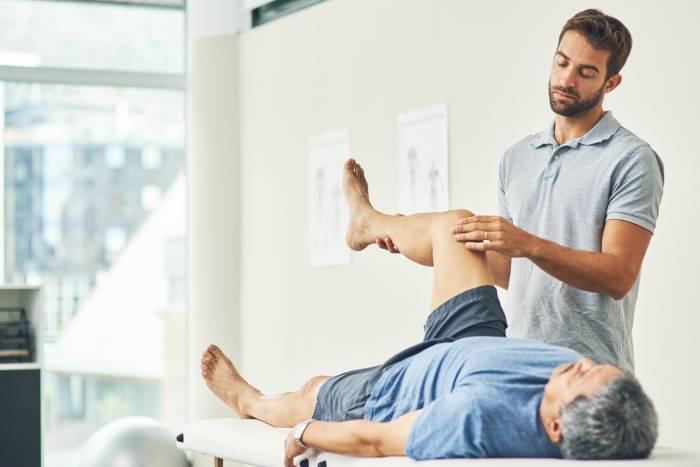 A physiotherapist lifting up the right leg of an elderly man lying on bed for treatment.