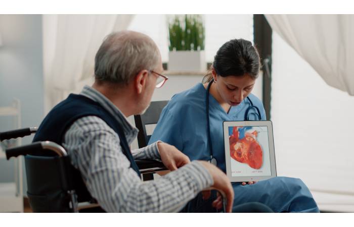 A cardiologist sitting on a chair holding a tablet displaying a heart figure explains it to an elderly man sitting on a wheel chair near her.